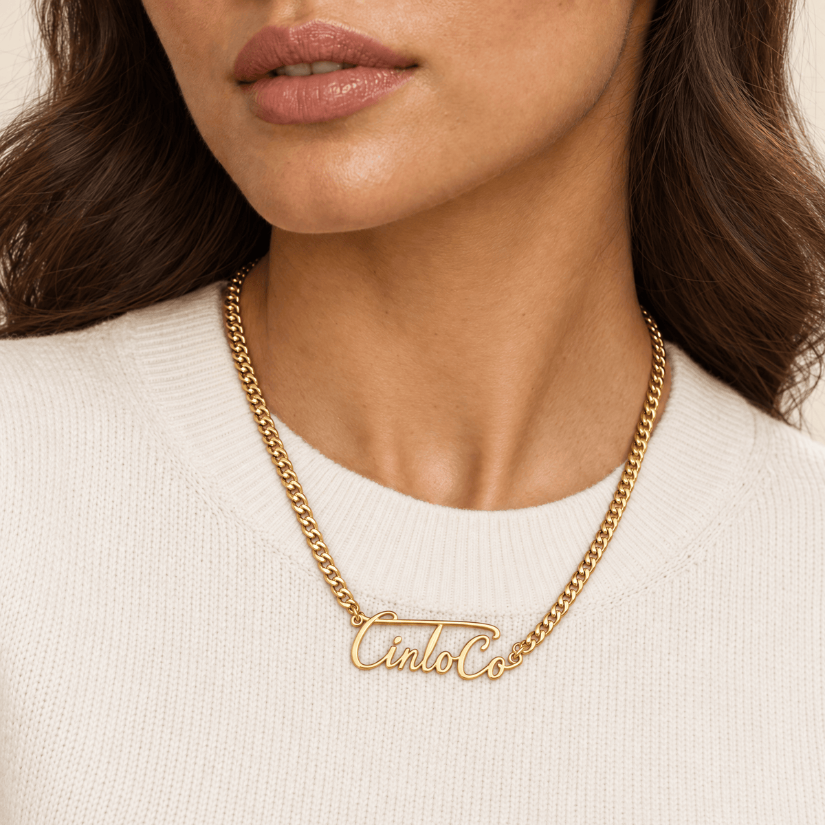 A close-up of a model wearing a gold chain necklace with a script nameplate pendant that reads 'Cinco Co'. The model is wearing a cream-colored knit top, and the background is a soft, warm neutral tone.
