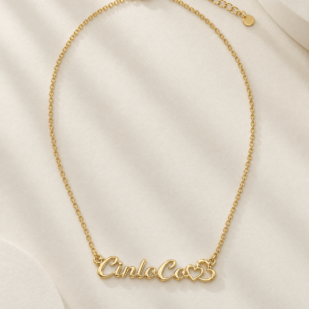 A delicate gold chain necklace with a personalized script nameplate reading 'Cinco de Mayo' in a cursive font, flanked by two small heart charms, displayed on a soft, draped cream fabric background.