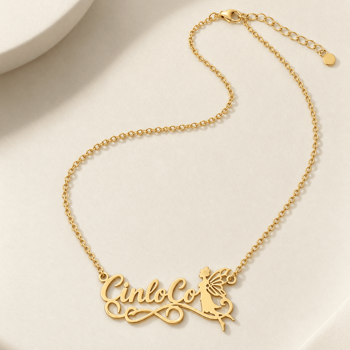 A gold nameplate necklace spelling 'CinloCo' with a small fairy silhouette detail, displayed on a clean, textured off-white surface.