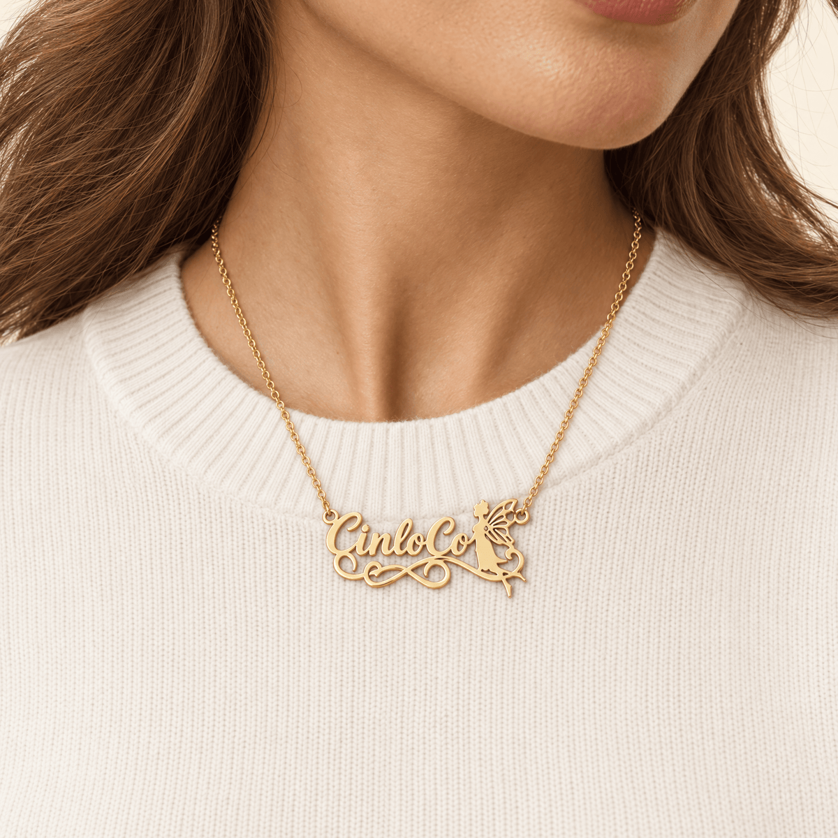 A close-up of a woman wearing a gold necklace with a custom script pendant spelling 'Cinco' and a small angel figure, paired with a cream-colored ribbed sweater.