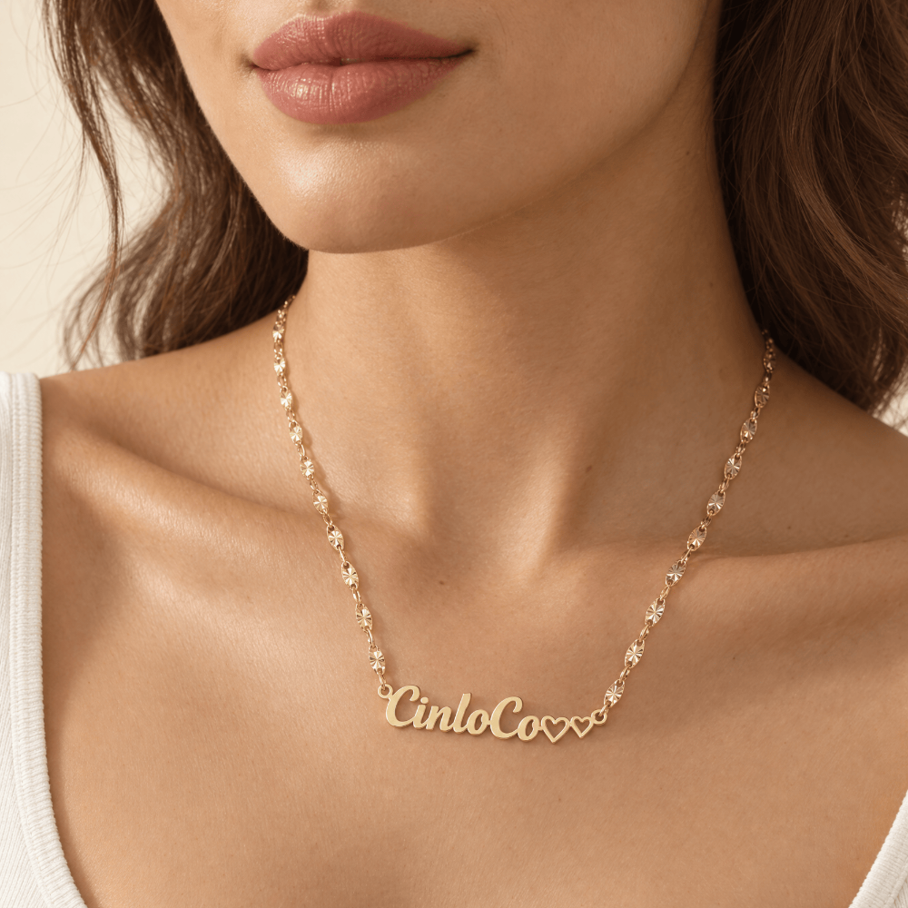 A close-up of a model wearing a delicate gold chain necklace with the name 'CinloCo' spelled out in script, accented by two small hearts at the end.