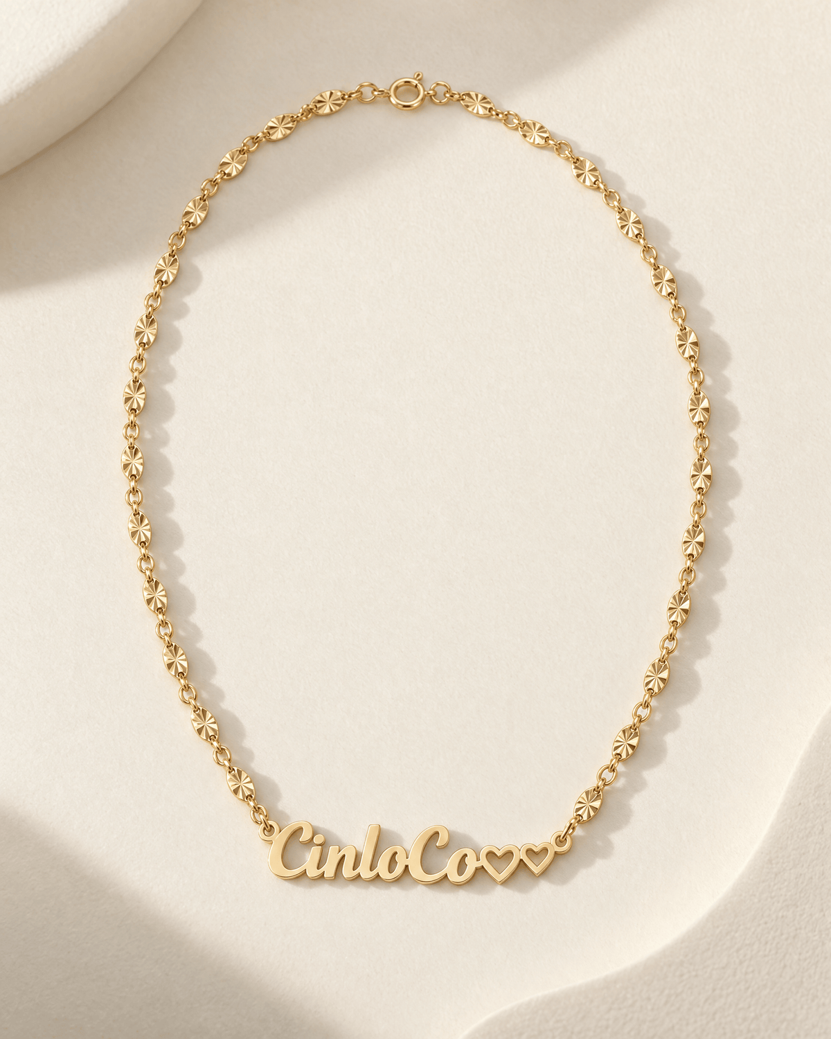 A delicate gold chain necklace with a personalized script nameplate reading 'CincoCo' and two small heart charms, displayed on a soft beige textured surface.