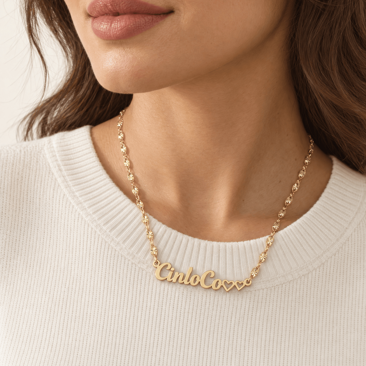 A close-up of a woman wearing a delicate gold chain necklace with a custom script nameplate reading 'Cinco Coxxx' in gold-tone letters, styled with three small hearts at the end.