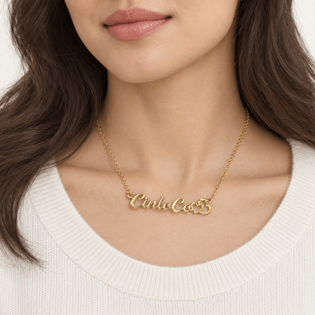 A close-up of a woman wearing a delicate gold nameplate necklace that reads 'Cinla Co. & S.' on a white ribbed knit top.
