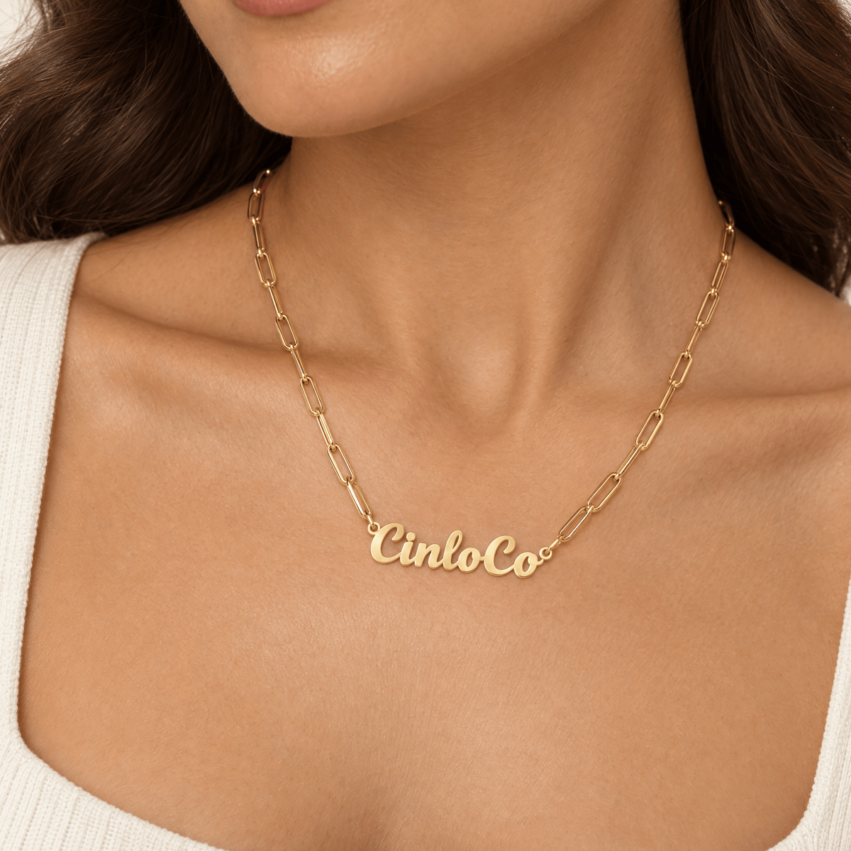 A close-up of a model wearing a gold nameplate necklace that reads 'CinloCo' on a delicate gold chain, paired with a white ribbed top.