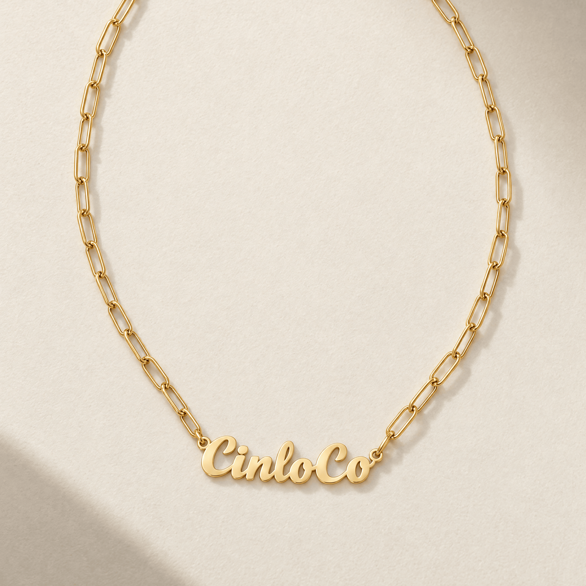 A gold paperclip chain necklace with a custom nameplate reading 'CinloCo' in cursive, displayed on a textured beige background with soft shadows.
