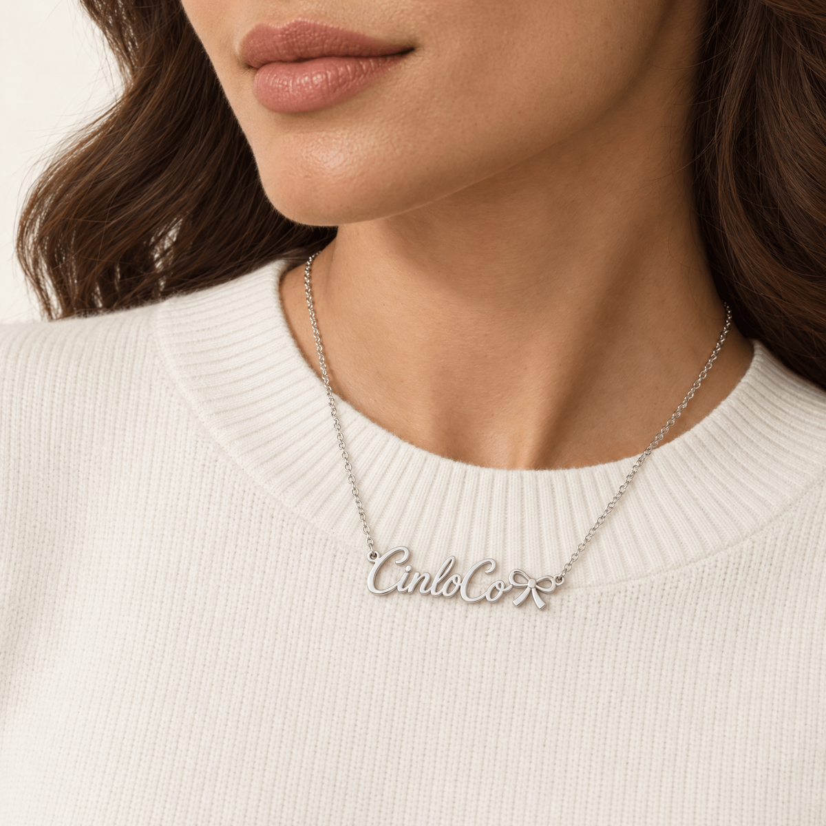 A close-up of a woman wearing a silver script nameplate necklace that reads 'CinkCoXX' over a white ribbed knit sweater.