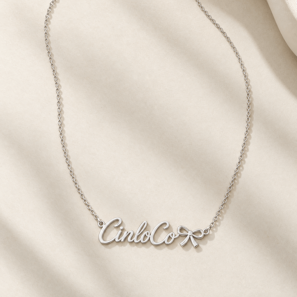 A silver name necklace with the script name 'Cinco' and a small bow charm, displayed on a draped cream-colored fabric background.
