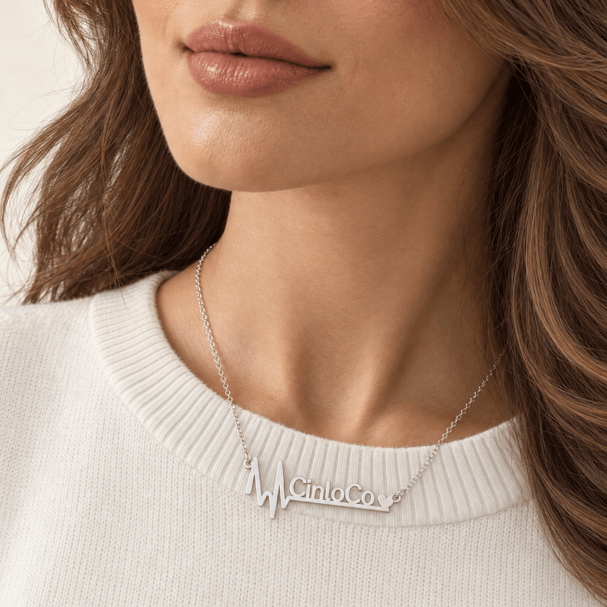 A close-up of a woman wearing a silver nameplate necklace that spells 'Mia Co.' with a heartbeat line motif, paired with a white ribbed knit top.