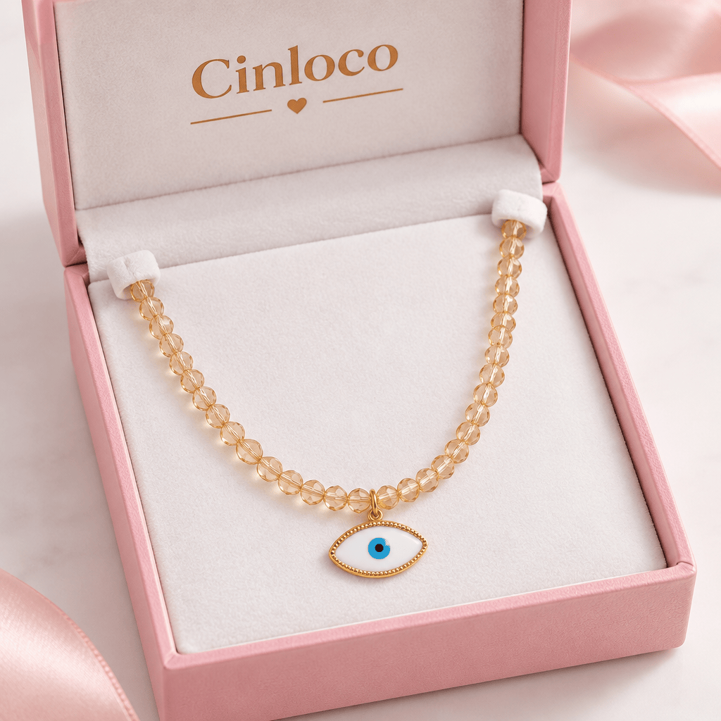 A beaded necklace with a gold-tone evil eye pendant displayed inside a pink jewelry box. The necklace features clear faceted beads and a gold-tone evil eye charm with a blue center.
