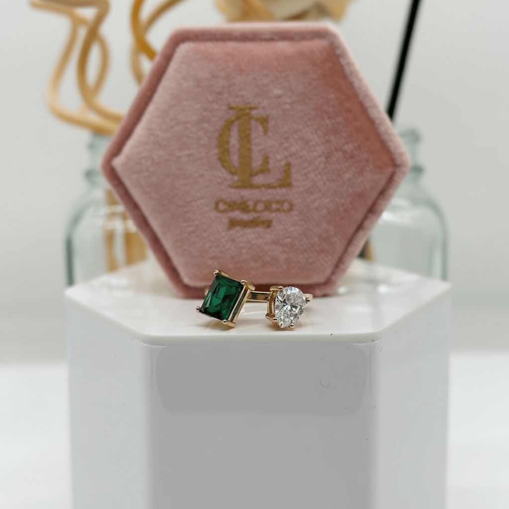 Pair of earrings with green gemstone and clear gemstone in front of a pink jewelry box with 'JC' logo.