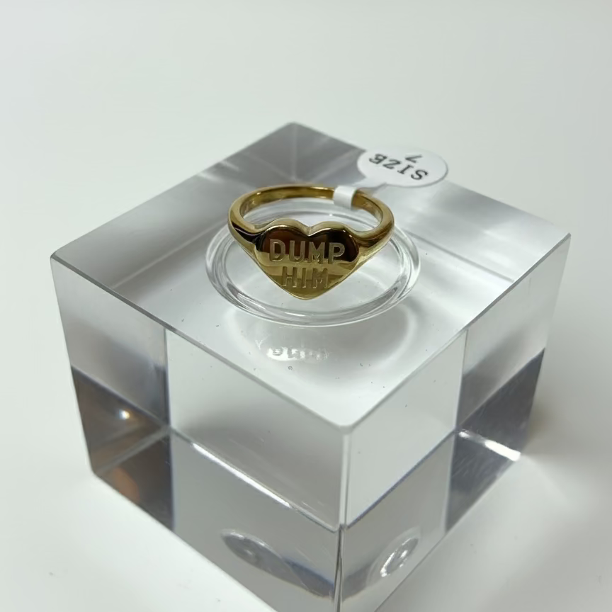 Gold ring with 'DUMP HIM' engraving on a clear stand against a white background